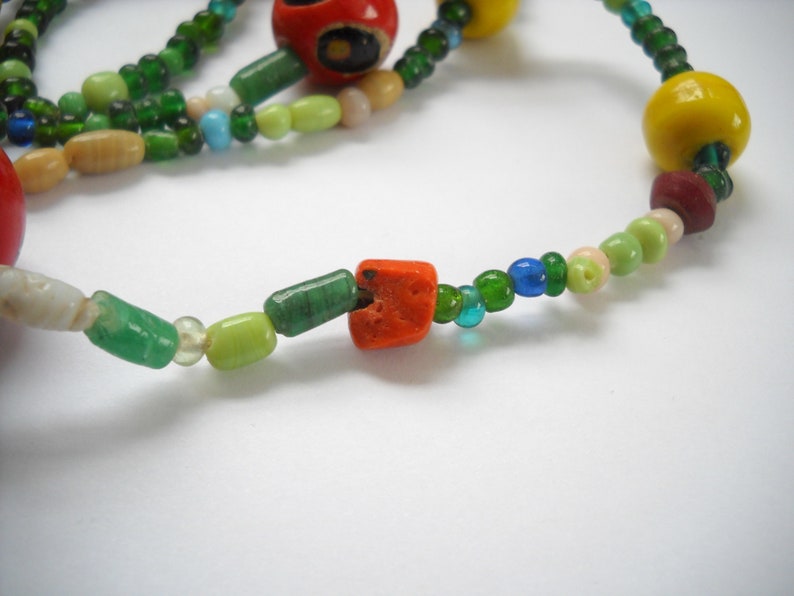 Op de afbeelding: Een kleurrijke kralenketting met groene, rode, gele en witte kralen. De ketting heeft verschillende vormen en maten kralen, waaronder ronde, vierkante en rechthoekige kralen.