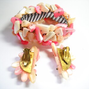 K&ouml;nnte beinhalten: Ein pinkfarbenes und wei&szlig;es Muschel-Armband und passende Ohrringe. Das Armband besteht aus einem Metallband, an dem pinkfarbene und wei&szlig;e Muschelteile befestigt sind. Die Ohrringe sind Clip-Ohrringe und haben ein &auml;hnliches Design wie das Armband.