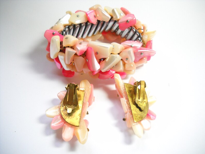 K&ouml;nnte beinhalten: Ein pinkfarbenes und wei&szlig;es Muschel-Armband und passende Ohrringe. Das Armband besteht aus einem Metallband, an dem pinkfarbene und wei&szlig;e Muschelteile befestigt sind. Die Ohrringe sind Clip-Ohrringe und haben ein &auml;hnliches Design wie das Armband.