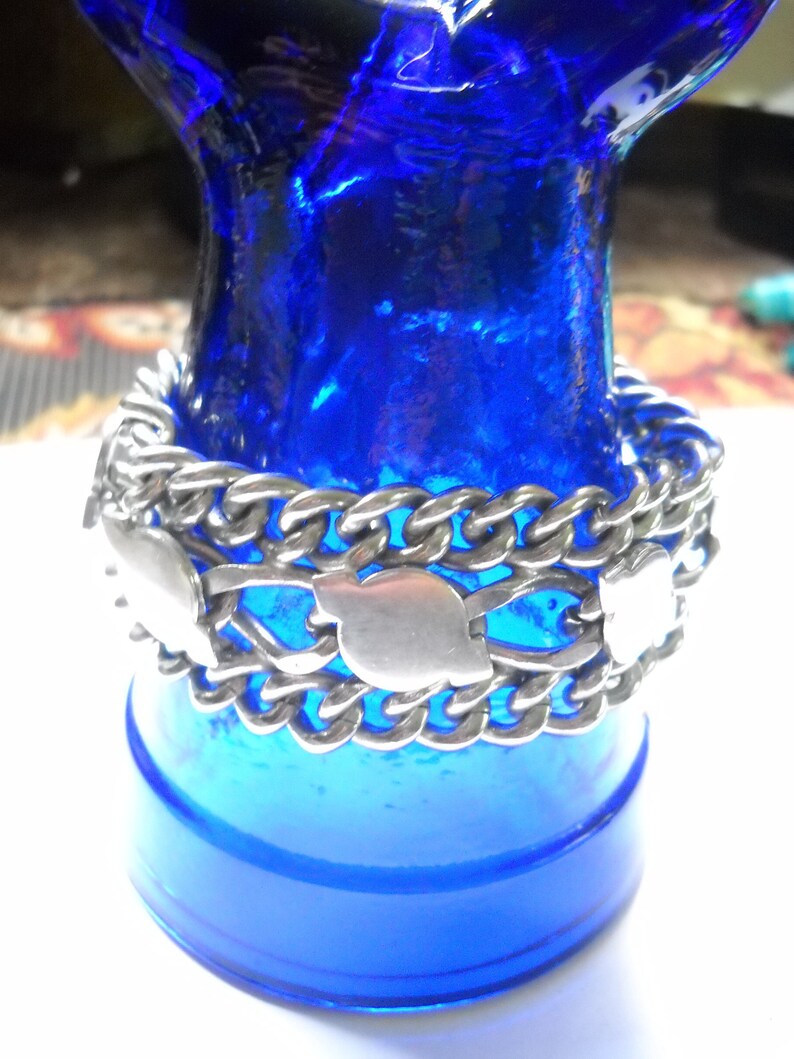 May include: A silver chain bracelet with a series of decorative silver heart-shaped charms. The bracelet is displayed on a blue glass bottle.