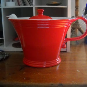 May include: A vibrant red ceramic teapot with a matching lid and a curved handle. The teapot has a white interior and a ridged design around the body. It sits on a wooden surface.