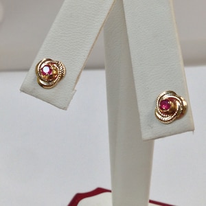 May include: A pair of gold earrings with a red gemstone in the center of each earring. The earrings are on a white display stand.