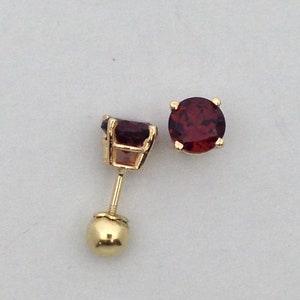 May include: A pair of gold stud earrings with red gemstones. The earrings have a round shape and are set in a prong setting.