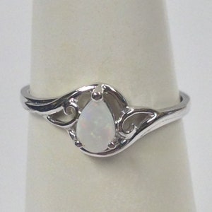 May include: A silver ring featuring a pear-shaped, white opal gemstone. The ring has a decorative, swirling design around the opal, with a polished finish. The ring is displayed on a white background.