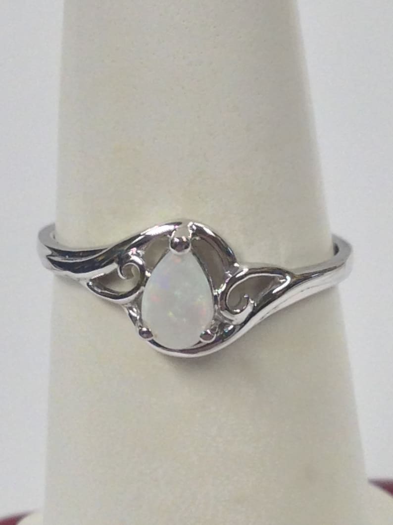 May include: A silver ring featuring a pear-shaped, white opal gemstone. The ring has a decorative, swirling design around the opal, with a polished finish. The ring is displayed on a white background.