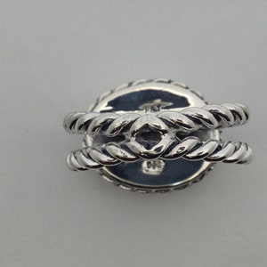 May include: Silver ring with a round, rope-like design. The ring has a raised center with a diamond-shaped opening.