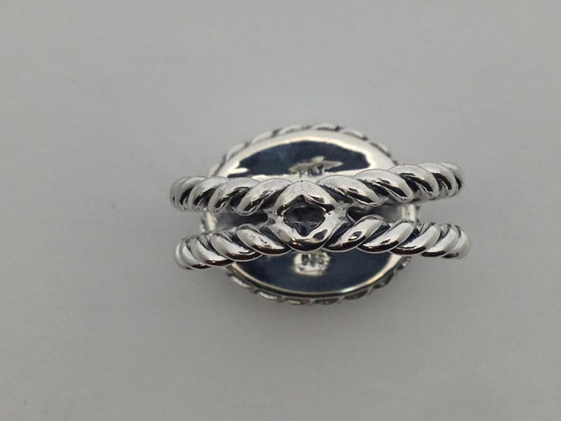 May include: Silver ring with a round, rope-like design. The ring has a raised center with a diamond-shaped opening.