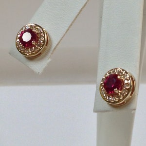 Puede incluir: Un par de pendientes de oro. Cada pendiente presenta una gema redonda de color rojo intenso rodeada por un halo de gemas más pequeñas y claras. Los pendientes se exhiben en tarjetas de exhibición de pendientes blancas.