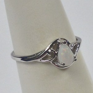 May include: A silver ring with an opal teardrop-shaped stone. The ring has an ornate, openwork design around the stone, with a polished finish. The opal has a milky white color.
