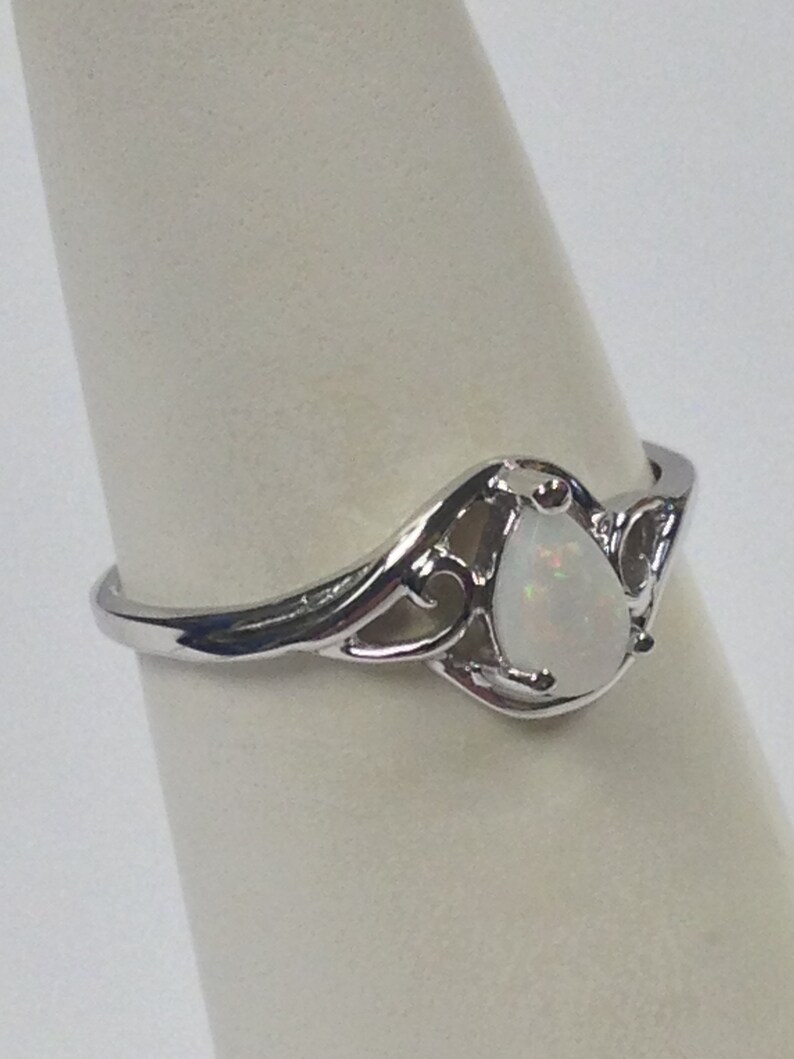 May include: A silver ring with an opal teardrop-shaped stone. The ring has an ornate, openwork design around the stone, with a polished finish. The opal has a milky white color.