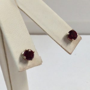 Op de afbeelding: Een paar gouden oorbel studs met rode edelstenen. De oorbellen zijn op een witte displaystandaard.