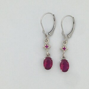 May include: A pair of silver dangle earrings with a red oval gemstone and a smaller red gemstone accent. The earrings have a leverback closure.
