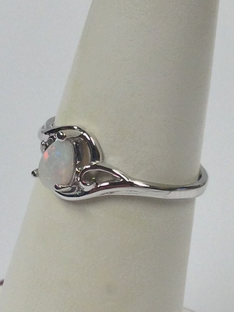 May include: A silver-toned ring featuring a pear-shaped, iridescent opal gemstone. The ring has a delicate, swirling design around the stone and along the band, showcasing a classic and elegant style. The opal displays a play of colors.