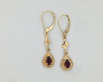 Könnte beinhalten: Ein Paar goldene Ohrringe mit einem tropfenförmigen roten Edelstein, der von kleineren weißen Edelsteinen umgeben ist. Die Ohrringe haben einen diamantförmigen Anhänger mit einem einzelnen weißen Edelstein.