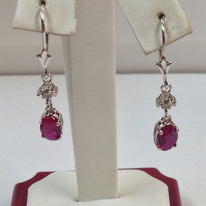 May include: A pair of silver dangle earrings with a red oval gemstone and small clear gemstones. The earrings are hanging from a white display stand.