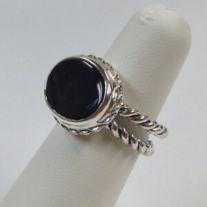 May include: A silver ring with a black oval gemstone set in a rope-like design. The ring is on a white ring display stand.