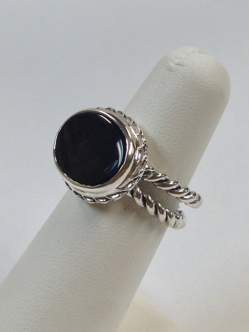 May include: A silver ring with a black oval gemstone set in a rope-like design. The ring is on a white ring display stand.