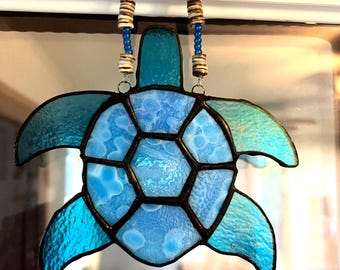 Könnte beinhalten: Eine Glasmalerei-Schildkröte mit einem blauen und hellblauen Design. Die Schildkröte hängt an einer weißen Wand mit einer braunen und blauen Perlenkette.