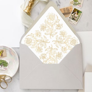 Può includere: Una busta bianca con un disegno floreale dorato sul davanti. La busta è appoggiata su una superficie di marmo con un biglietto bianco, un sigillo di cera dorato e un piattino con francobolli e una farfalla.