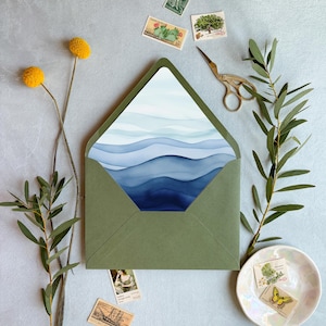 Può includere: Una busta verde oliva con il lembo aperto che rivela un inserto di cancelleria con un design ad acquerello di onde oceaniche blu stratificate. La scena include due fiori gialli billy ball, un paio di forbici dorate e diversi francobolli vintage.