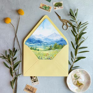 May include: A pale yellow envelope containing a card with a watercolour landscape painting of blue mountains, green hills, and a wildflower meadow. The scene includes vintage postage stamps, a brass crane-shaped scissor, and dried yellow flowers.