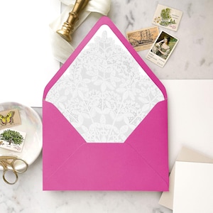 Puede incluir: Un sobre rosa brillante con un forro blanco con estampado floral. El sobre está abierto, revelando el intrincado diseño. Sellos postales vintage y un sello de cera dorado son visibles cerca, lo que sugiere suministros de papelería o invitación.