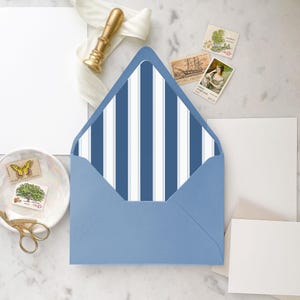 Puede incluir: Un sobre azul claro con un interior a rayas azules y blancas. El sobre está abierto, revelando el patrón de rayas. Varios sellos postales vintage, un sello de cera dorado y tarjetas en blanco están dispuestos alrededor del sobre, lo que sugiere artículos de papelería o invitación.