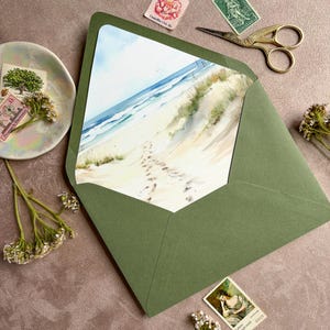 May include: An olive green envelope containing a card with a watercolor beach scene featuring sand dunes, ocean waves, and footprints in the sand. The envelope is surrounded by vintage postage stamps, gold-toned scissors, and small white flowers.