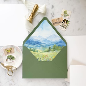 May include: A green envelope with a watercolor illustration of a mountain landscape with a blue sky and green fields. The envelope is decorated with a white ribbon and a gold wax seal.