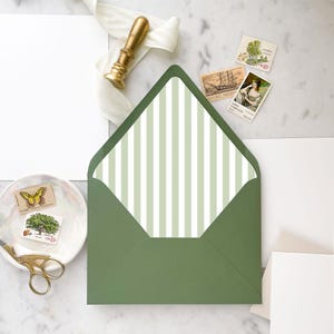May include: A green envelope with a striped interior, alongside stationery items. A gold wax seal, stamps, and a small dish with scissors are visible. The envelope's interior has vertical stripes in light green and white.