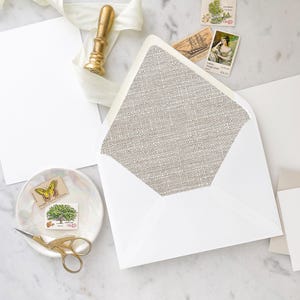 May include: A white envelope with a linen-textured inner flap. The envelope is lying on a white marble surface with a gold stamp, a white ribbon, and a few stamps.