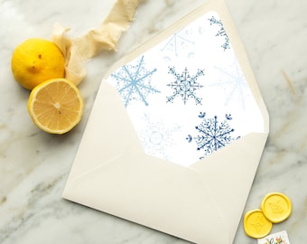 May include: An open cream-colored envelope with a snowflake pattern in shades of blue. A lemon and a half-lemon are on the left. Two yellow wax seals and a small floral stamp are on the right. The envelope is on a marble surface.