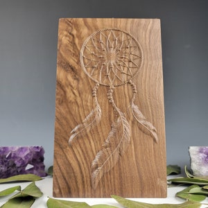 May include: A wooden wall hanging featuring a carved dreamcatcher design. The dreamcatcher has a circular frame with a web-like pattern and feathers hanging from the bottom. The wood has a natural brown colour with a grain pattern.