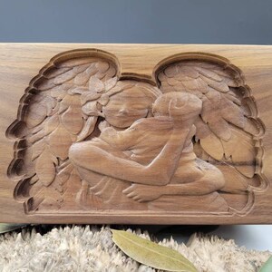 May include: A wooden box featuring a carved image of an angel with wings holding a child. The box is crafted from dark wood and has a smooth finish.