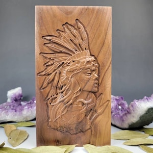 May include: A wooden wall plaque featuring an intricate carving of a woman's face adorned with a headdress of feathers. The wood is a rich brown colour.