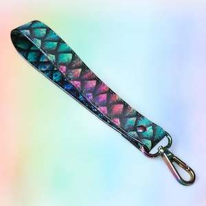 May include: A wristlet keychain strap featuring a repeating diamond scale pattern in vibrant shades of teal, magenta, and black. The strap is secured with a metallic rivet and includes a rainbow-finished metal lobster clasp at the end.