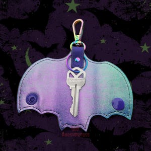 May include: A bat-shaped key holder in iridescent purple and teal, with a silver key attached. The holder has a snap closure and a rainbow-colored clasp. The text "Odd Awesomeness" is at the bottom.