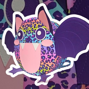 May include: A purple and pink bat-shaped backpack with a colourful leopard print design. The backpack has a large, toothy grin and big eyes. The text "Odd Awesomeness" is printed on the bottom of the backpack.