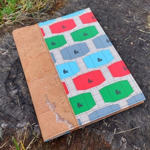 Puede incluir: Un cuaderno con respaldo de corcho y un patrón geométrico colorido. El patrón presenta formas rojas, azules, verdes y blancas con un diseño repetitivo. El cuaderno tiene un respaldo de corcho marrón.