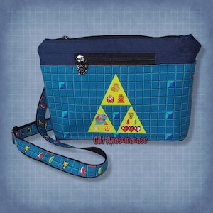 Op de afbeelding: Een blauwe schoudertas met een pixelachtig rasterpatroon en een grote gele triforce-afbeelding met diverse videogamepersonages en hartpictogrammen. De tas heeft een donkerblauw bovenpaneel, een ritsvak met een schildhanger en een bijpassende riem.