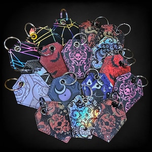 May include: A collection of 15 colorful keychains with various designs, including lace, skulls, and geometric patterns. The keychains are made of metal and leather and have a variety of colors, including black, white, red, blue, and green.