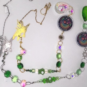 May include: A collection of handmade necklaces with various charms and beads. One necklace features a silver fairy frog charm, while another has a gold fairy charm. Other necklaces include green beads, iridescent accents, and circular pendants with the text "IT'S A BEAUTIFUL DAY TO SMASH THE PATRIARCHY".