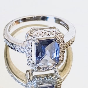 Puede incluir: Un anillo de plata con una gran piedra azul de forma cuadrada rodeada de diamantes más pequeños. El anillo tiene una banda que está engastada con diamantes más pequeños.
