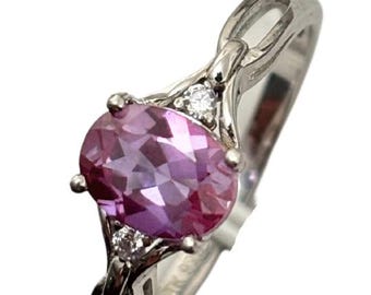 Può includere: Un anello d'argento con una pietra ovale rosa e due pietre più piccole trasparenti. L'anello ha una fascia delicata e intrecciata.