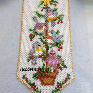 Può includere: Un'appendice da parete a forma di albero di Natale in perline bianche con cinque uccelli colorati e una stella dorata in cima. L'albero è in un vaso marrone con foglie verdi e bacche rosse. Il testo "HobbyPerle" è visibile nella parte inferiore dell'immagine.