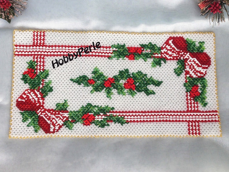 Op de afbeelding: Een witte, met kralen bezette kersttafelrunner met rode en groene hulst- en strikdesigns. De runner heeft een gouden kralenrand en de tekst "HobbyPerle" is in het midden geborduurd.