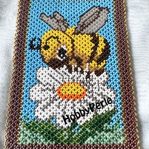 Pode incluir: Uma imagem de contas de uma abelha em uma margarida. A abelha é amarela e preta com olhos castanhos. A margarida é branca com um centro amarelo. A imagem é emoldurada em contas marrons e douradas. O texto "HobbyPerle" está no canto inferior direito.