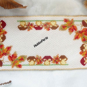 Può includere: Un sottobicchiere quadrato di perline bianche con un bordo di perline dorate. Il centro del sottobicchiere è bianco e il bordo presenta un disegno di foglie rosse, arancioni, gialle e verdi e funghi marroni.