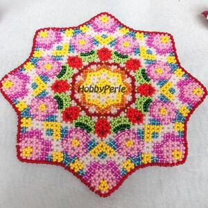May include: A colorful beaded mandala with a starburst shape. The center of the mandala is a circle with a red, yellow, and green design. The mandala is surrounded by a ring of pink, yellow, and blue beads.
