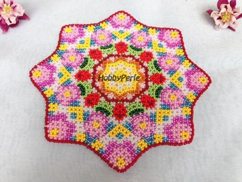 May include: A colorful beaded mandala with a starburst shape. The center of the mandala is a circle with a red, yellow, and green design. The mandala is surrounded by a ring of pink, yellow, and blue beads.
